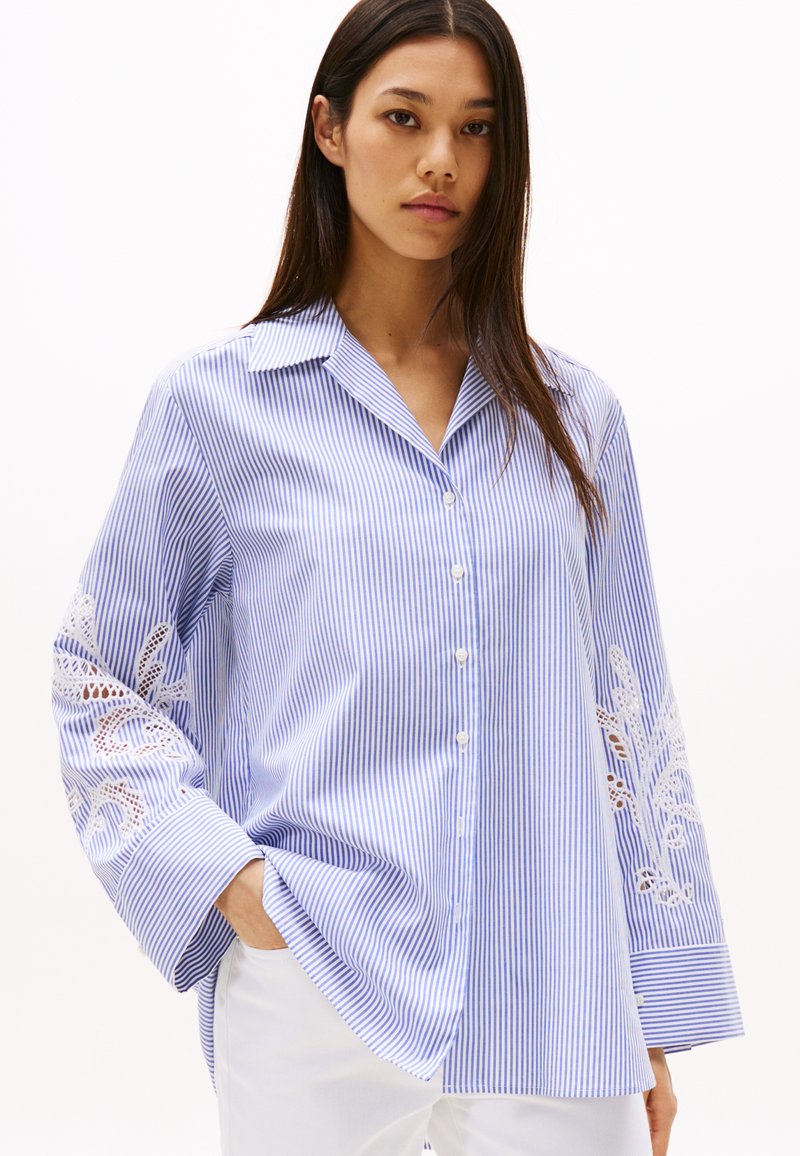 Camicia a righe blu e bianche con maniche lunghe e un dettaglio floreale in pizzo sulla manica sinistra; presenta un colletto classico e chiusura con bottoni.