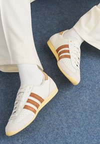 White sneakers with brown suede stripes, leather and textile materials, perforated toe, and yellow rubber sole. Worn with white socks.