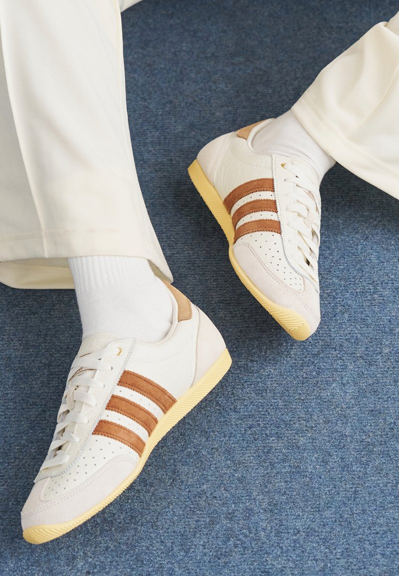 White sneakers with brown suede stripes, leather and textile materials, perforated toe, and yellow rubber sole. Worn with white socks.