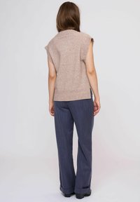 Beige gebreide mouwloze trui met geribbelde zoom, gecombineerd met donkerblauwe pantalon met streep. De broek heeft een wijde pijp met manchet bij de enkels.