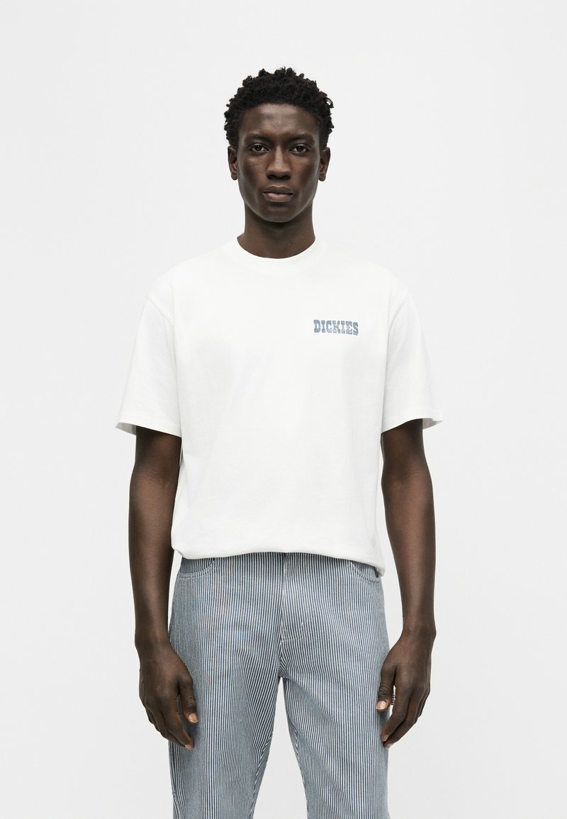 Man wearing white short-sleeve t-shirt with "DICKIES" text and light blue and white pinstriped pants standing against a plain background.