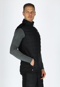 Schwarze gepolsterte Weste mit hohem Kragen, Reißverschluss und Seitentaschen. Über einem grauen Langarmshirt getragen; minimalistisches Design mit schlanker Erscheinung.