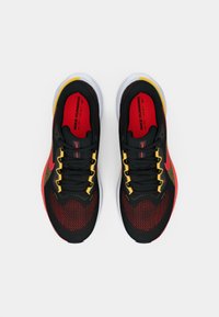 Zapatillas deportivas negras con acentos rojos y amarillos. Parte superior de malla, suela blanca gruesa y cordones de contraste. Destacado logotipo rojo de Nike en el lateral.