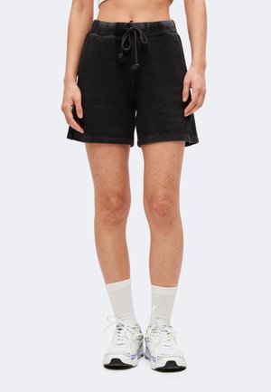 Person iført sorte ribstrikkede shorts med snøre, hvide crew-sokker og hvide sportssko står mod en ensartet lys baggrund.