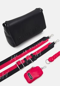 Musta nahkainen olkalaukku, jossa on kohokuvioitu logo. Laukkua on varustettu kahdella hihnalla: toisessa on punamusta teksti, ja toisessa punainen punottu hihna. Lisäksi mukana on pieni punainen pussi.