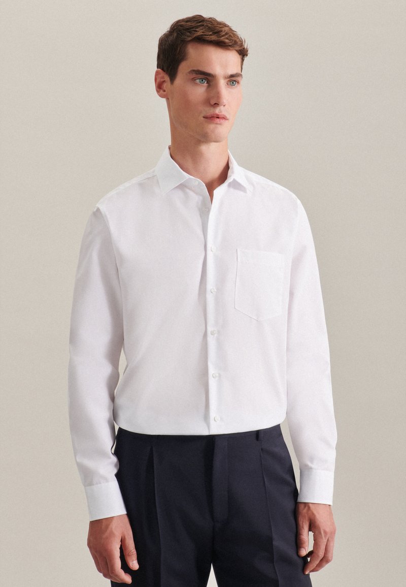 Witte lange mouwen shirt met een klassieke kraag, voorzak en knopenlijst; gecombineerd met marineblauwe broek, gladde stoftextuur.