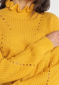 Pull en tricot jaune avec un col haut volanté, des accents ajourés et des détails de poignets côtelés. Texture douce avec une coupe ample.