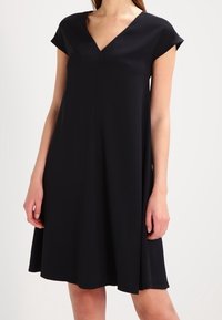 Černé šaty s krátkým rukávem, V-výstřihem, volným střihem a hladkým materiálem, které sahají nad kolena. Minimalistický design bez vzorů.