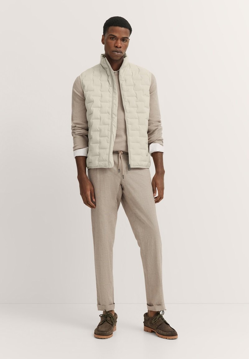 Hellgraue gesteppte Weste über einem beigen Rippensweater und beigen Hosen, kombiniert mit dunkelbraunen Schuhen, im entspannten Schnitt und minimalistischen Design.