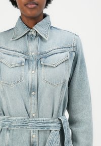 Light blue denim shirt dress with a collar, two chest pockets, and a belted waist. Features silver button closures and visible stitching details.