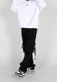 Hvid hættetrøje med et lille sort logo, parret med sorte cargo-bukser med lommer og stropper, suppleret med sort og hvidt sneakers.