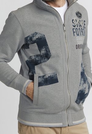 Hombre con chaqueta gris claro con cremallera, con un gran número 24 azul marino desgastado en el frente y texto en la parte superior derecha del pecho, con las manos en los bolsillos.