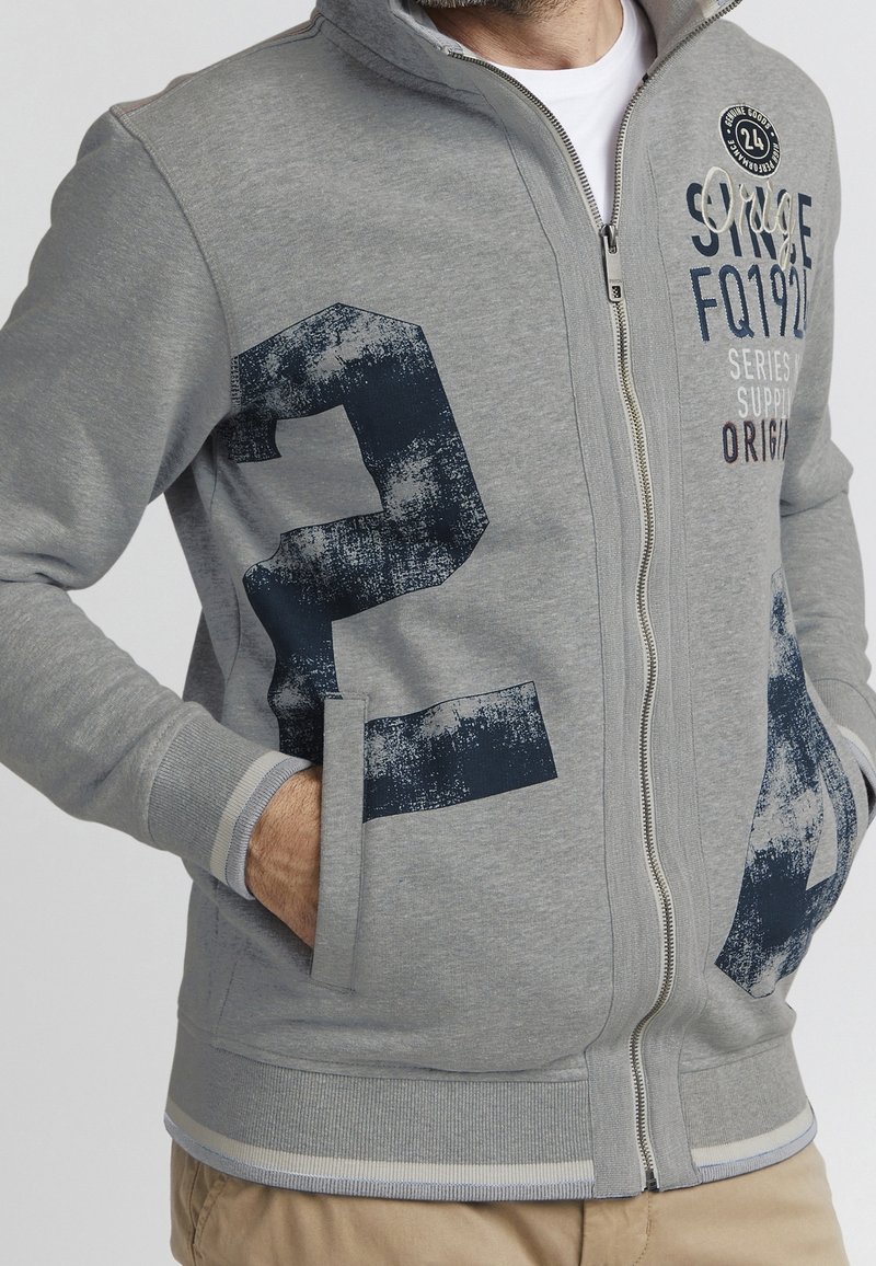 Homme portant une veste zippée gris clair avec un grand numéro 24 bleu marine usé sur le devant et un texte sur la poitrine droite supérieure, les mains dans les poches.