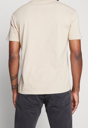 Homme portant un t-shirt beige uni à manches courtes et un jean gris foncé, debout dos à la caméra.