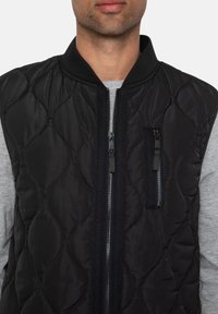 Gilet imbottito nero con colletto alto, chiusura con zip sul davanti e maniche lunghe grigie, dotato di tasca laterale con zip e tessuto testurizzato.
