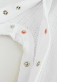 Weißer Stoff mit feiner Struktur und dezenten Mustern. Entlang des Kragens befinden sich drei farbige Druckknöpfe: silber, gold und rosé.