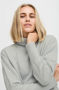 Pull à col roulé gris clair, à côtes, avec des manches longues, présentant une texture douce et une coupe décontractée, couvrant partiellement les bras du mannequin.