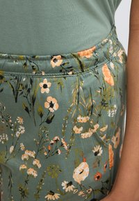 Πράσινο ύφασμα με λουλουδάτο σχέδιο που περιλαμβάνει πορτοκαλί και  λευκά λουλούδια. Λάστιχο στη μέση με απαλή υφή, κατάλληλο για καθημερινή χρήση.