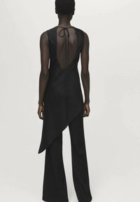 Zwarte jumpsuit met een vloeiende, asymmetrische zoom en open achterkant. Voorzien van een striksluiting bij de nek en een wijduitlopende pijp. Soepele textuur.