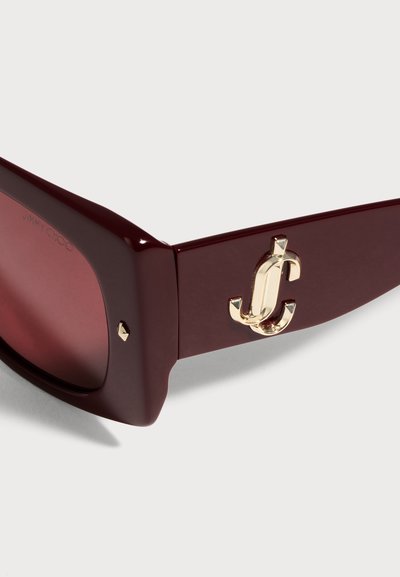 Gros plan sur des lunettes de soleil Jimmy Choo bordeaux, avec des verres teintés foncés et un logo doré sur la branche, sur un fond clair.