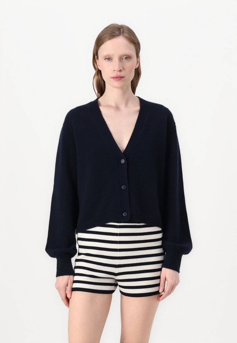 Marinefarvet cropped cardigan med V-hals, lange ærmer og fire knapper. Båret over sort-hvide stribede højtaljede shorts.