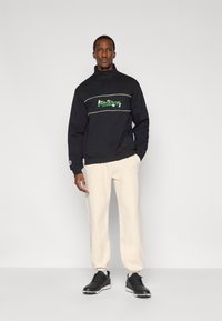 Μαύρο φούτερ με φερμουάρ στο μισό, με κεντητό πράσινο κείμενο, σε συνδυασμό με εκρού φόρμες και μαύρα αθλητικά παπούτσια, με άνετη εφαρμογή και ριμπ μανσέτες.