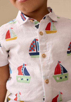 Enfant portant une chemise blanche à boutons avec des motifs de voiliers colorés en rouge, bleu, vert et jaune.