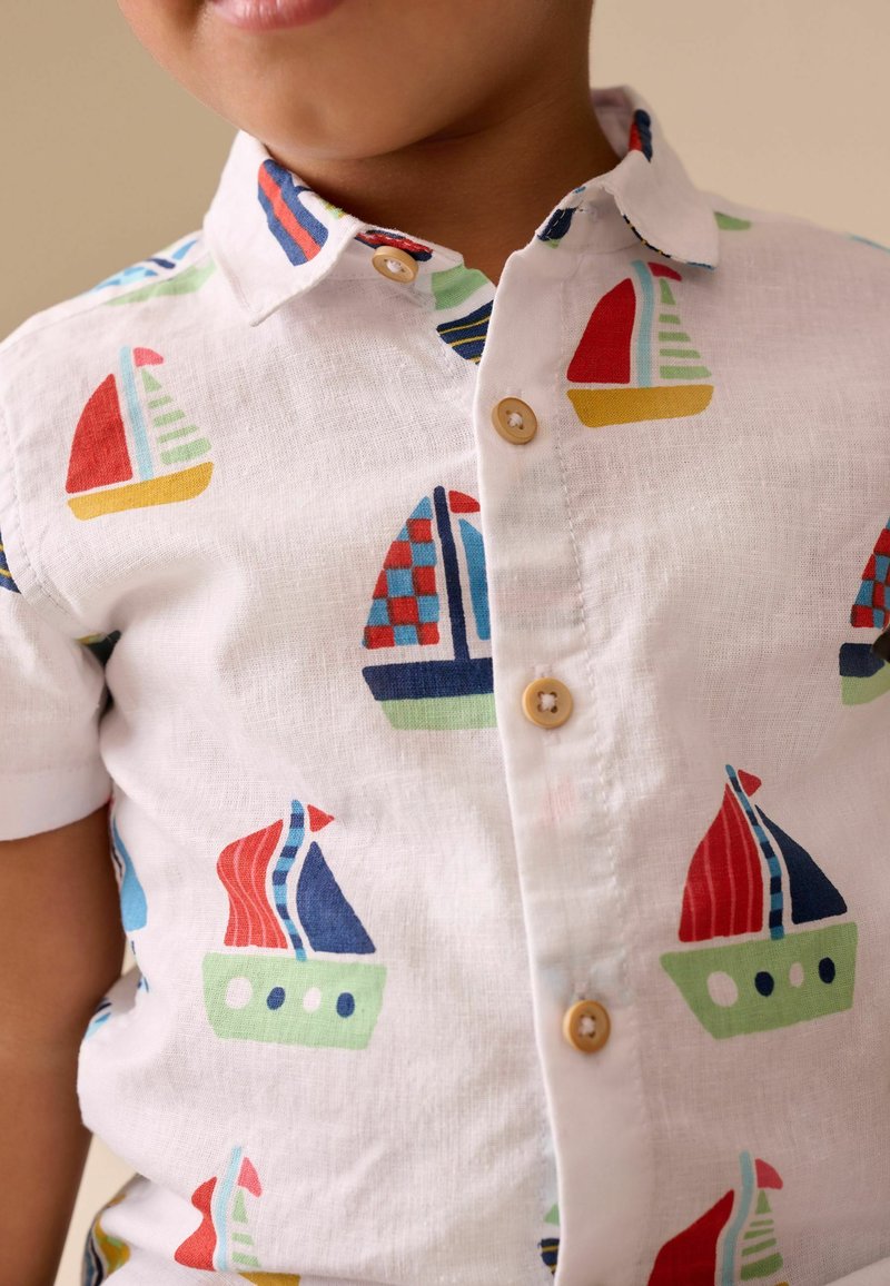Enfant portant une chemise blanche à boutons avec des motifs de voiliers colorés en rouge, bleu, vert et jaune.