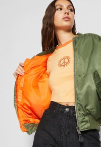 Maslinasto zeleni bomber jakna razrezana da otkrije svijetlu narančastu podlogu; narančasta majica s kružnim grafičkim prikazom; nosi se s crnim farmerkama.