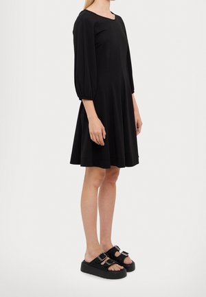 Robe noire à encolure carrée, manches trois-quarts bouffantes et jupe évasée. Associée à des sandales compensées noires avec des boucles.