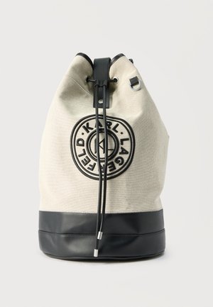 Beige og svart bøtteveske med snøring, med sirkulært Karl Lagerfeld-logo på fronten og svarte snorer med metalltupper.