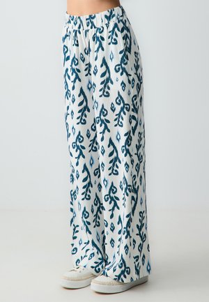 Wijdvallende broek in witte stof met een blauw botanisch patroon. Elastische tailleband, luchtige textuur, gecombineerd met witte schoenen.