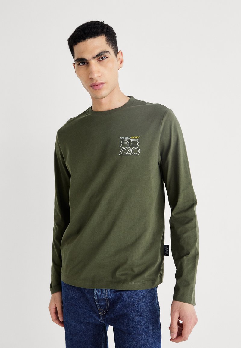 Olīvzaļa, garām piedurknēm, kokvilnas krekls ar mazu balto un dzelteno grafisko logotipu uz krūts, saskaņots ar ziliem džinsu biksēm.