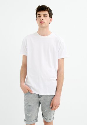 Joven de pie con una mano en el bolsillo, vistiendo una camiseta blanca lisa y pantalones cortos de mezclilla gris claro remangados sobre un fondo blanco.