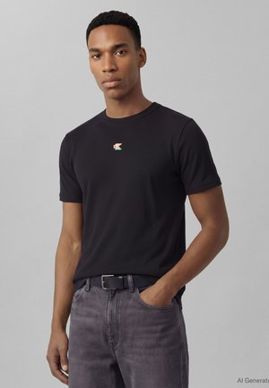 Άνδρας που φοράει μαύρο κοντομάνικο πουκάμισο με μικρό πολύχρωμο λογότυπο "CK", γκρι τζιν και μαύρη ζώνη, στέκεται με το ένα χέρι στην τσέπη.