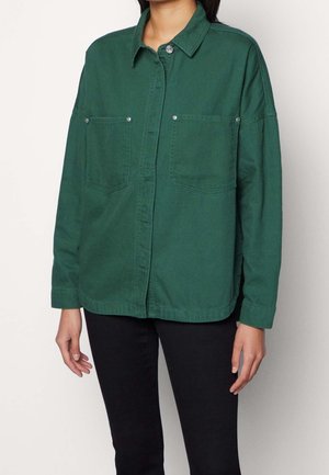 Persoon draagt een groene blouse met lange mouwen en twee borstzakken en een zwarte broek, staand tegen een effen achtergrond.