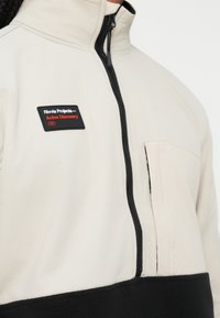 Jasnobeżowy polarowy sweter z czarnymi akcentami, wysokim kołnierzem i zamkiem błyskawicznym do połowy. Posiada naszywkę z logo na piersi oraz kieszeń z przodu.