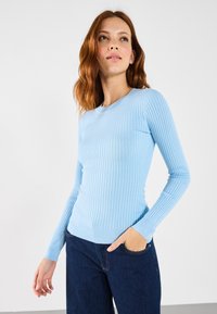 Lichtblauwe geribbelde langemouwen top met een ronde halslijn en een getailleerd ontwerp. De stof heeft een gladde textuur; het model draagt donkere spijkerbroek.