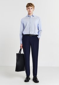Giovane uomo che indossa una camicia azzurro pallido con bottoni, pantaloni blu navy, cintura nera, scarpe nere, mentre tiene una grande borsa a tracolla nera.