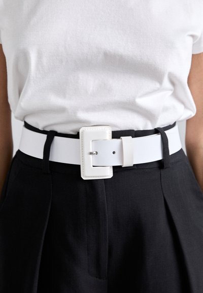 Curea din piele ecologică albă, cu un cataramă dreptunghiulară, purtată peste pantaloni negri. Curea are o textură netedă și un design elegant, modern.