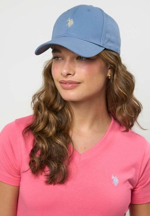 Tânără cu păr castaniu ondulat, purtând o șapcă bleu deschis și un tricou roz cu decolteu în V, cu un mic logo brodat pe piept.