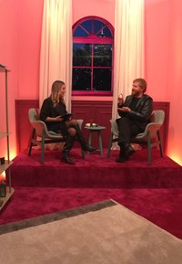 To personer sitter i moderne stoler på en scene med dyp rødt teppe. Et rundt bord holder skjønnhetsprodukter. Rosa vegger og et stort vindu er synlige.