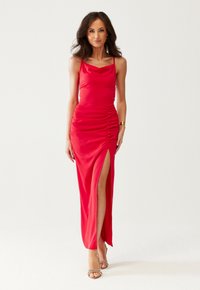 Vrouw in een rode mouwloze gerimpelde jurk met een hoge split tot boven de dijen en huidskleurige gespsandalen, staande tegen een eenvoudige witte achtergrond.