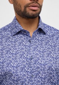 Camicia a motivi floreali blu con collo a giro, maniche corte e tessuto morbido. Presenta bottoni scuri in contrasto con il motivo.