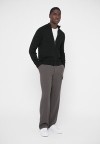 Camisola preta com fecho de correr, gola e punhos canelados, combinada com calças cinzentas largas e sapatilhas brancas, criando um visual casual.