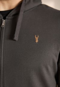 Tumši pelēkā krāsā izgatavota kapuce ar rāvējslēdzēju, kas izgatavota no mīksta materiāla, ar nelielu, gaiši brūnu izšūtu briedi uz krūtīm.