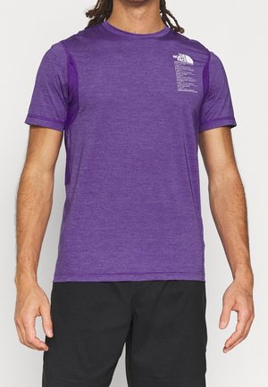 Man draagt een paarse korte mouwen T-shirt van The North Face en zwarte korte broek, staat met ontspannen armen langs zijn zij.
