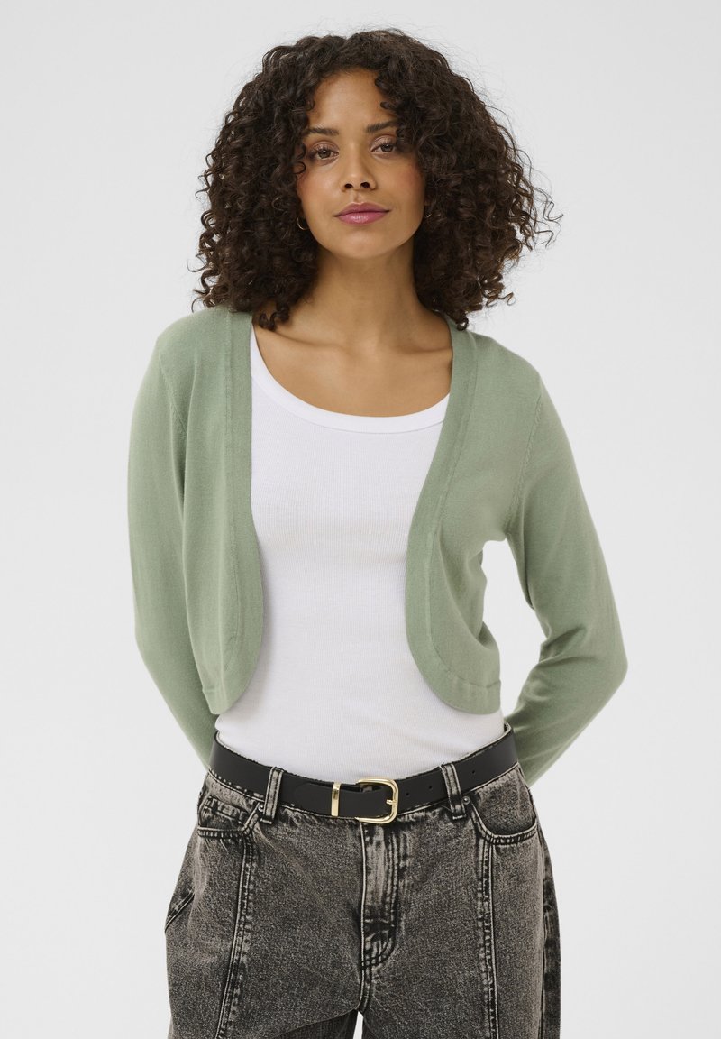 Groen, lichtgewicht vest met een ronde, open voorkant en lange mouwen, gedragen over een wit overhemd, gecombineerd met hooggetailleerde, gestructureerde zwarte jeans.