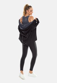 Sort lynlåstrøje, grå tanktop og sorte leggings. Modellen har hvide sneakers på, og tøjet viser en glat stoftekstur og tætsiddende design.