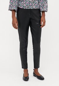 Calças de couro pretas com um design ajustado e textura lisa, combinadas com sapatos planos pretos, contra um fundo branco liso.